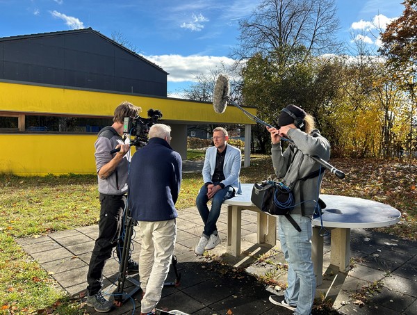 Ein Mann sitzt im Garten auf einer Tischtennisplattte und wird vom Fernsehteam interviewt